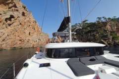 2022 Lagoon 46 catamaran sailing near rocky cliffs under clear blue sky.